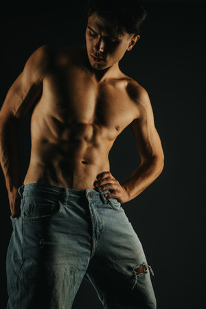 Young shirtless man showcasing defined abs in ripped jeans in dramatic lighting