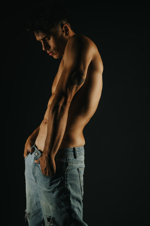 Shirtless man in jeans posing against a dark background highlighting muscle definition and tattoosの写真素材