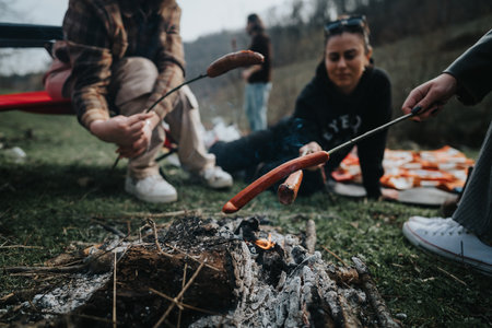 Friends enjoying a casual outdoor barbecue with roasted sausages over a campfireの写真素材