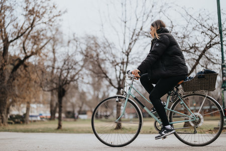 Side view of a woman in casual attire cycling along a park path with trees in the background during an overcast day.の写真素材