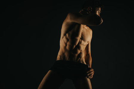 Fit male showing muscular torso and abs under dramatic lighting in a dark studioの写真素材