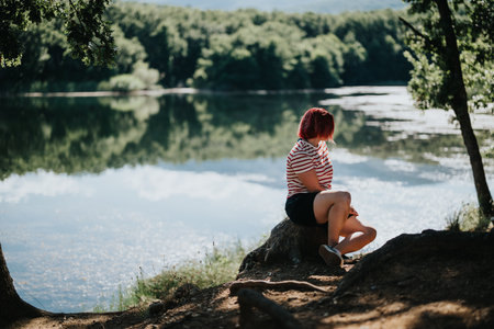 Woman relaxing by the lake in summer, enjoying a peaceful moment in nature surrounded by trees and waterの写真素材