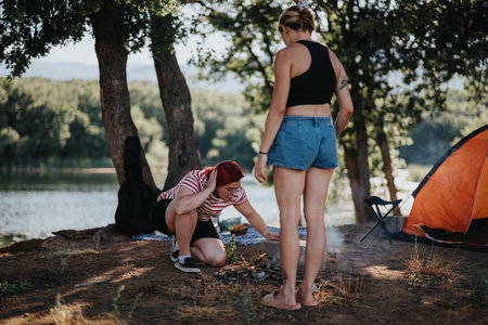 Two friends preparing campfire during mountain picnic near lake, setting up barbeque for outdoor adventureの写真素材
