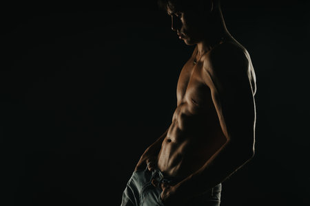 Fit muscular man posing shirtless in dim lighting showcasing abs and muscles