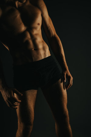 Fit male showing muscular torso, abs, and arms in dark lighting for bodybuilding and fitnessの写真素材