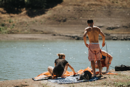 Group of friends setting up a tent beside a peaceful lake on a sunny dayの写真素材