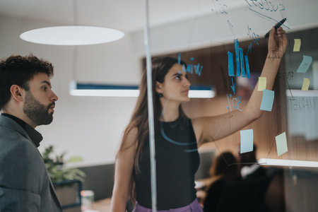 European male and Indian female business colleagues brainstorming with sticky notes on a glass wall in modern office setting.の写真素材