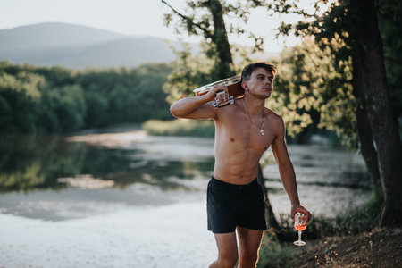Young man enjoying nature with a glass of wine on a summer day by a peaceful lakeの写真素材