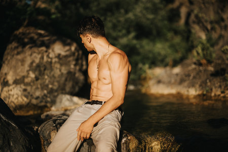 Shirtless man sitting on rocks outdoors near water in natural sunlightの写真素材