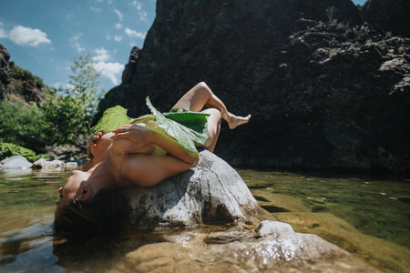 Woman relaxing on rock near river holding large leaf in natural setting with clear blue skyの写真素材