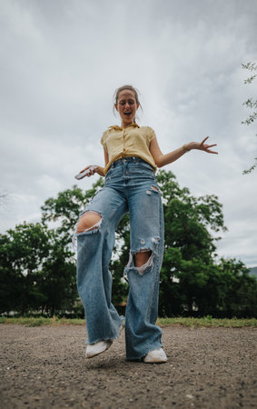 Young woman in torn jeans joyfully dancing outdoors on a cloudy dayの写真素材