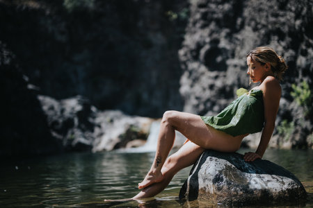 Woman in leaf dress relaxing on rock by serene river in nature settingの写真素材