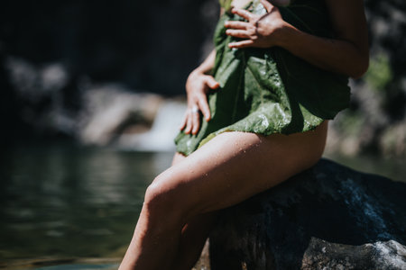 Woman in nature covering herself with a large leaf while sitting by a serene lakeの写真素材