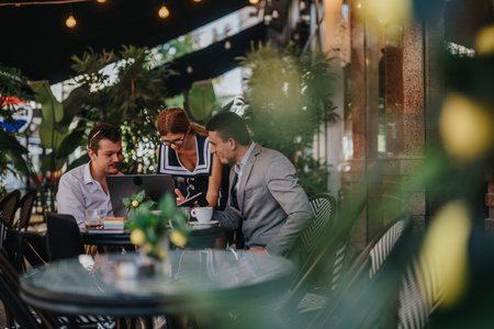 Business team collaborating at an outdoor cafe with a laptop and coffee, surrounded by lush greenery and warm lighting,の写真素材