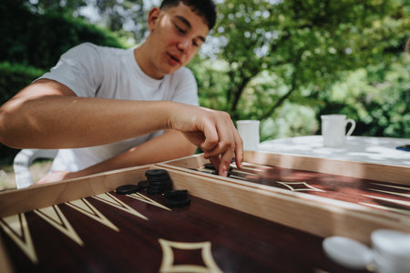 Young man enjoys a sunny day while playing a game of backgammon outdoors, surrounded by nature with a relaxing atmosphere, highlighting leisure time and social interaction.の写真素材