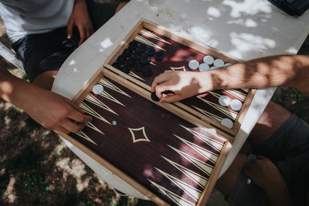 Outdoor backgammon game with two people enjoying a relaxing board game on a sunny day at a table under dappled shade from trees in the backgroundの写真素材