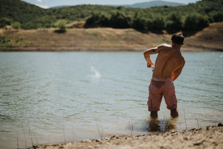 Man skipping stones in a serene lake setting surrounded by lush green hills on a sunny dayの写真素材