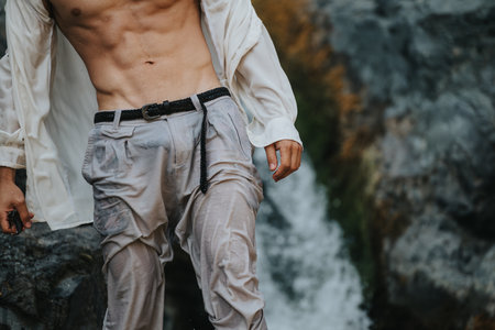 Man with muscular torso in wet clothes standing at the edge of a waterfall