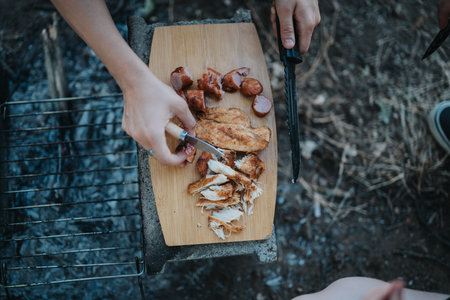 Friends enjoying a barbeque picnic in nature, grilling and slicing delicious meatの写真素材