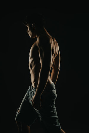 Fit young man with muscular physique posing in dark studio lighting wearing ripped denim shortsの写真素材