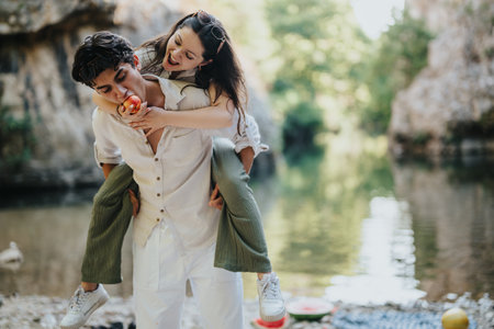 Joyful young couple enjoying a playful piggyback ride by a serene riverside during a sunny day outing in nature, sharing laughter and fresh fruit in a peaceful outdoor setting.の写真素材