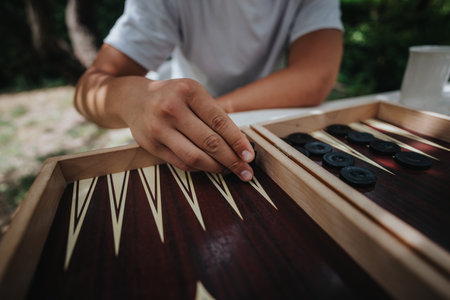 Close-up of a person playing backgammon outdoors on a sunny day, focusing on strategic game play with wooden board and black checkersの写真素材