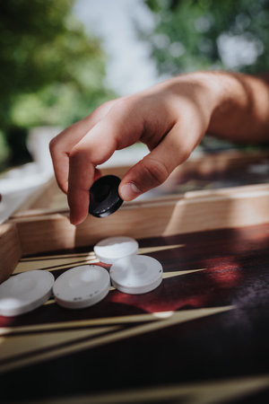 Close-up of a hand playing backgammon outdoors, capturing a strategic move under sunlight and shadow
