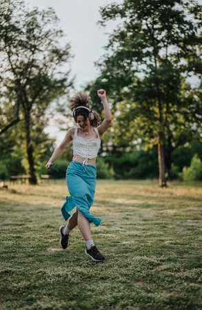 Young woman dancing joyfully in a sunny park with headphonesの写真素材