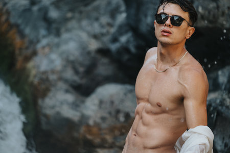 Young muscular man posing shirtless by the rocky beach in sunglassesの写真素材