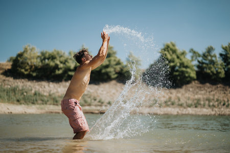 Young man enjoying a sunny day outdoors, splashing water in a lakeの写真素材