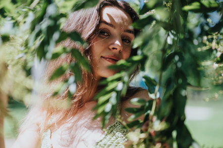 Serene young woman smiling through green foliage on a sunny day in the parkの写真素材
