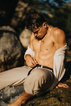 Shirtless man sitting by a river with wet hair and casual clothing on a sunny dayの写真素材