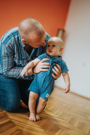 Father helping baby take first steps on wooden floor at home