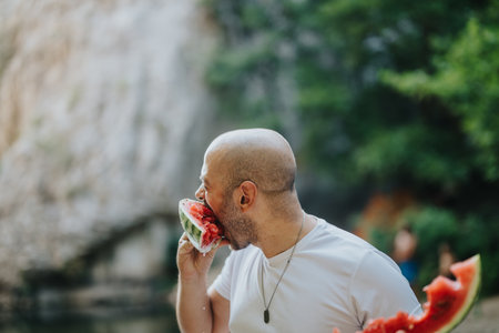 Man enjoying fresh watermelon on a summer day by the water. Refreshing and healthy snack outdoors, capturing the essence of summer joy and relaxation with lush green natural background.の写真素材