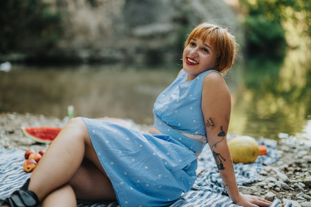 Joyful woman relaxing by the river on a summer day, surrounded by nature, enjoying a picnic with fresh fruit.の写真素材