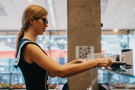 Young woman in a cafe wearing sunglasses, picking up coffee from a countertop with a calm expressionの写真素材