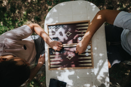 Two people playing backgammon outdoors on a sunny day, enjoying a friendly game under dappled...
