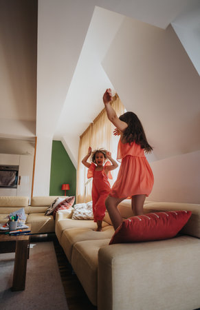 Young girls joyfully jumping on a cozy living room couchの写真素材