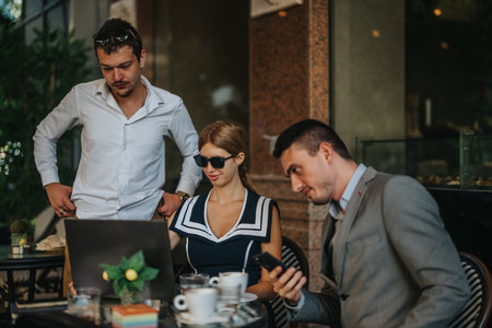 Team of businesspeople working together outdoors at a cafe, collaborating on a project with a laptop and smart phone, discussing strategies during a sunny day in a relaxed settingの写真素材