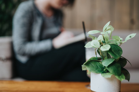 Woman writing in a notebook with a potted plant in the foreground, blurred backgroundの写真素材