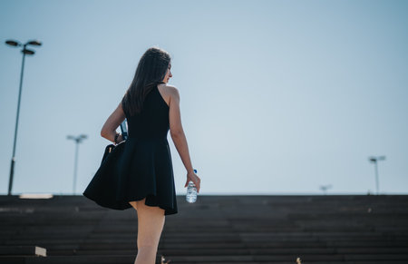Rear view of a woman in a black dress holding a water bottle walking up stairs outdoorsの写真素材