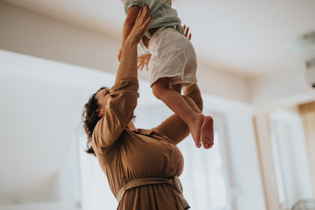 Mother joyfully lifting her young child in the air at homeの写真素材