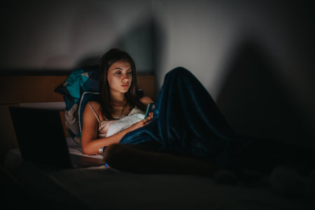 Young woman using smart phone in bed with laptop at night