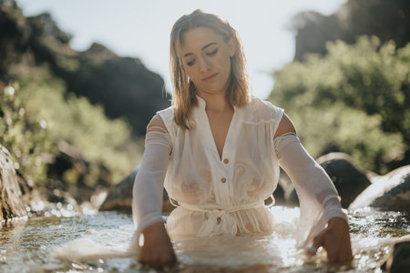 Serene woman in a flowing white dress sitting in a tranquil river settingの写真素材