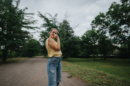 Young woman in casual outfit standing on a park path surrounded by lush green trees