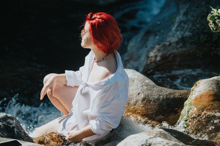 Red-haired woman relaxing by a river in a white shirt, enjoying the natureの写真素材