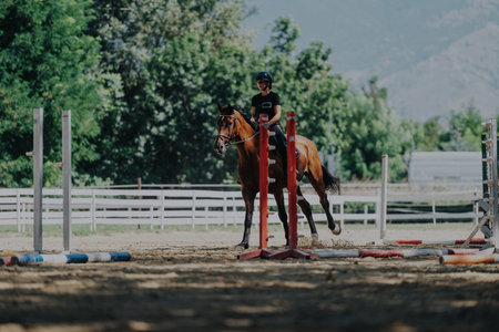 Young rider practicing horse jumping in sunny outdoor equestrian arena