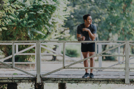 A young man stands casually on a wooden bridge, surrounded by lush greenery in a tranquil park...