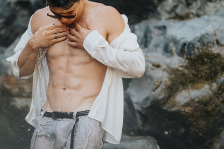 Fitness-focused man with damp shirt revealing toned abs under gentle waterfall by rocky landscapeの写真素材