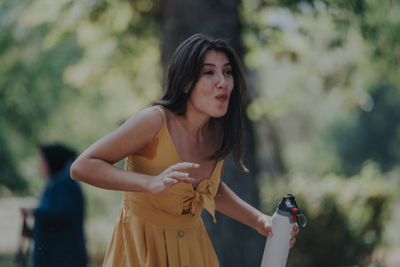 A cheerful woman in a yellow dress laughs while holding a water bottle in a sunlit park, embodying carefree joy and outdoor leisure with friends.の写真素材